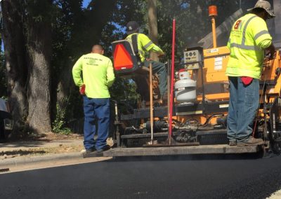 Workers in high-visibility clothing operate paving equipment while laying asphalt on a Charlotte, NC road.