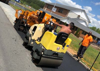 Construction workers in NC are busy with asphalt paving, using heavy machinery on a sunny day. In the background, a building and an American flag add to the scene.