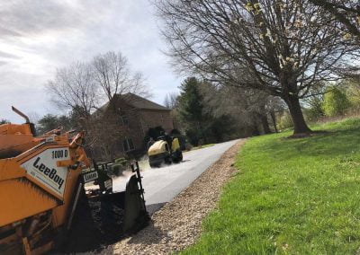 A worker operates a yellow asphalt roller on a newly paved road beside a house, as an asphalt paver machine works nearby, completing the latest project in North Carolina.