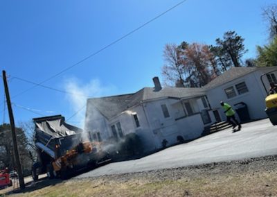 Construction workers paving a driveway in front of a white house on a sunny day in North Carolina, with one worker in a high-visibility vest.