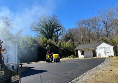 A person operates a yellow road roller to perform asphalt paving on a driveway near a small white building with trees in the background in North Carolina.