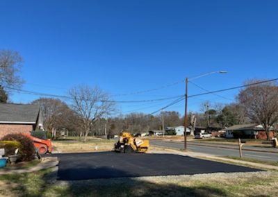 A paving machine smooths freshly laid asphalt on a residential street under the clear blue NC sky, with houses in the background.