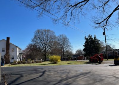 Neighborhood scene with trees and houses under a clear blue sky, a red tractor on the right, and freshly paved asphalt road in the foreground, capturing the charm of North Carolina.