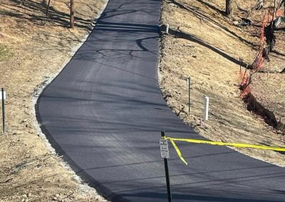 Newly asphalt paved driveway leading up to a house on a hill, with surrounding trees and construction tape.