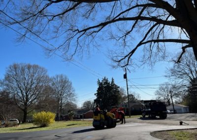 Workers using a steamroller for asphalt paving on a residential street in NC, with trees and houses visible in the background on a sunny day.