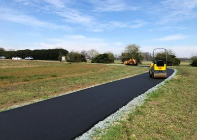 A small steamroller smooths a newly paved asphalt path in a grassy field in Charlotte, NC, with a blue sky and some cars in the background.