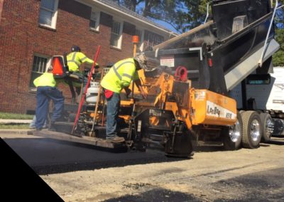 drivewat paving Construction workers in yellow vests using machinery to pave an asphalt road near a red brick building and white truck, ensuring the paving project in NC moves smoothly.