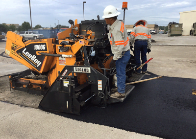 Construction workers operate heavy machinery to lay asphalt on a road under a clear sky in Charlotte, North Carolina.