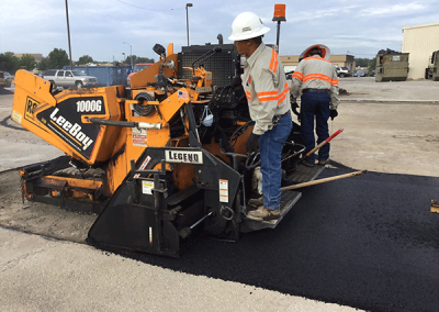 Two construction workers operate an asphalt paving machine on a road under a cloudy sky in Charlotte, NC.