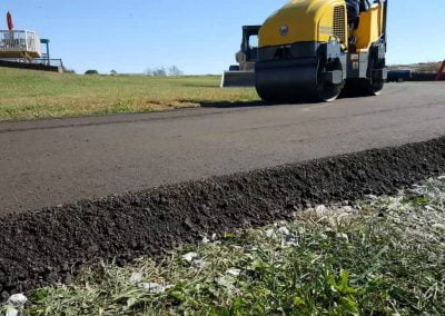 A yellow asphalt roller smoothing a newly paved road surrounded by grass on a sunny day in Charlotte.