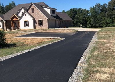 A newly paved asphalt driveway leads to a large, modern house with a mix of brick and wood siding, surrounded by trees.