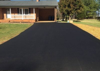 A newly paved asphalt driveway leads to a charming brick house with a porch railing, surrounded by the lush greenery typical of North Carolina.