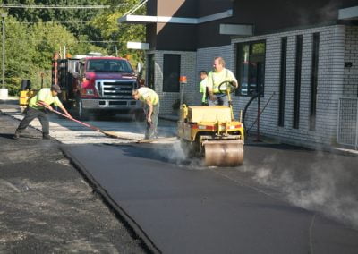 Construction workers in NC are busy resurfacing a road beside a building, using a steamroller and other equipment for asphalt paving.