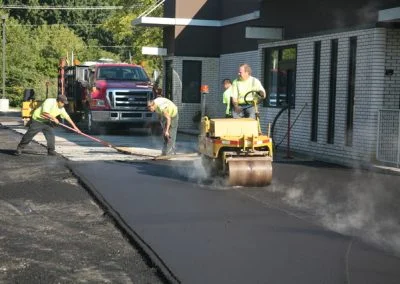 Workers in NC are busy with asphalt paving, using a steamroller and other equipment, right in front of a building.