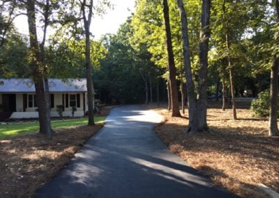 A paved path winding through trees with sunlight filtering through. A house is partially visible on the left, nestled in the serene landscape of NC.