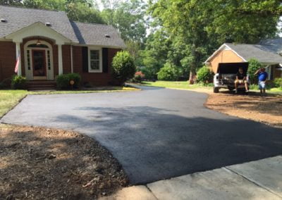 Freshly paved asphalt driveway in front of a brick home, with two people standing near the back of a truck on the right.