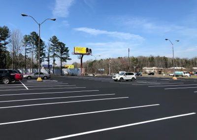 Nearly empty parking lot with just a few cars parked under streetlights on the fresh asphalt, and a large billboard in the background.