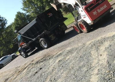 A Bobcat loader and truck work on a gravel road next to a grassy area under the sunny North Carolina sky.