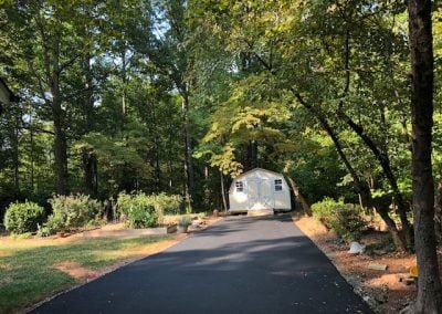 A paved driveway in Charlotte, NC, leads to a white shed surrounded by lush green trees and a garden on a sunny day.