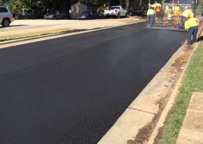 Workers in North Carolina are paving a road with asphalt on a sunny day, using heavy machinery, surrounded by trees and parked vehicles.