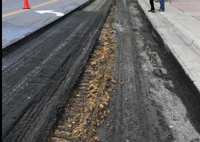 Workers repaving a city street with asphalt paving machinery and equipment, surrounded by traffic cones, pedestrians on the sidewalk in Charlotte, NC.
