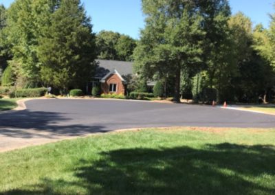 A brick house with a dark gray roof is surrounded by trees and located at the end of a cul-de-sac in Charlotte, North Carolina, featuring freshly laid asphalt paving.