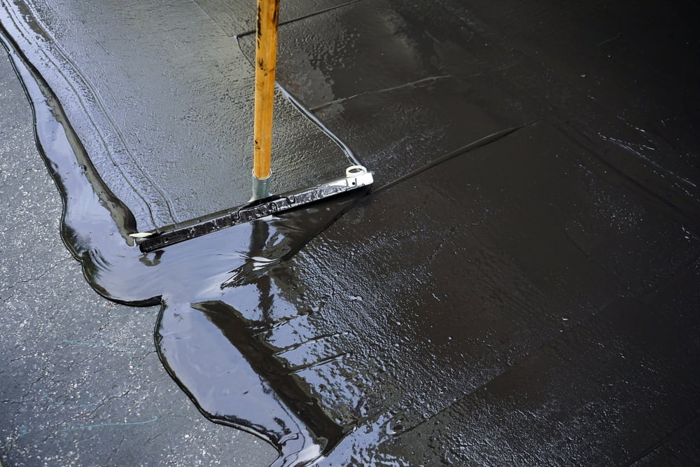A person in Charlotte, North Carolina, methodically spreading black, liquid asphalt on a driveway using a squeegee tool.