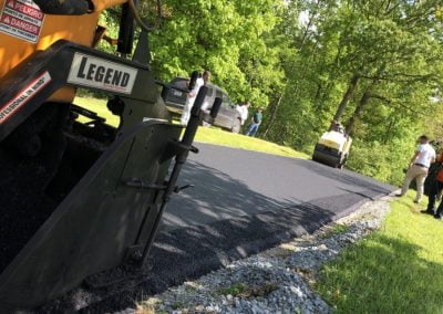 Asphalt Paving Cost Workers in Charlotte, NC, are paving a road with fresh asphalt on a sunny day, surrounded by green trees and grass.