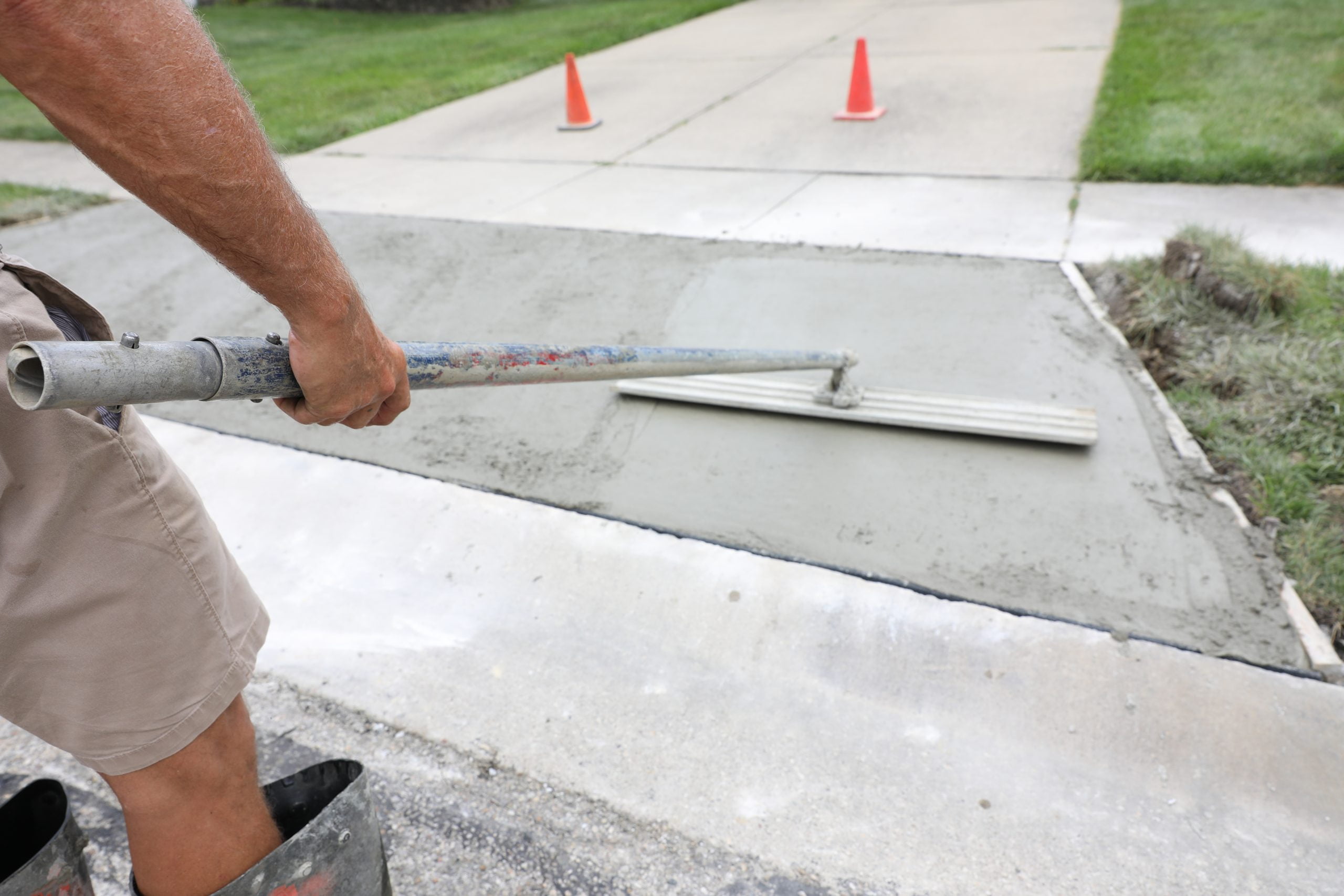 concrete work is being performed at a home in a urban neighborho Worker smoothing wet concrete on sidewalk with a long-handled tool in Charlotte, North Carolina, orange cones in background.