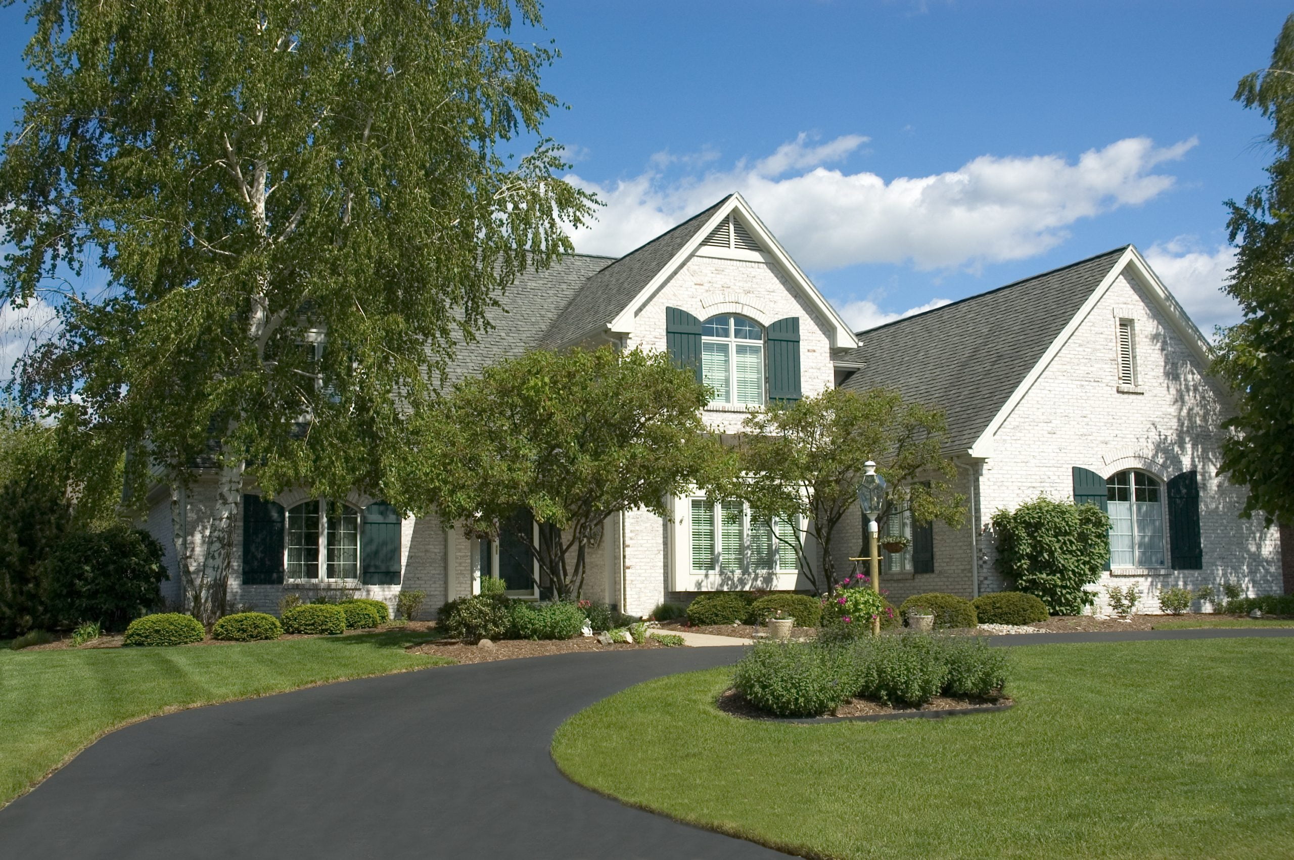 A large white-brick house with green shutters is nestled in the lush greenery of Charlotte, NC, featuring a well-manicured lawn and a winding, beautifully paved driveway.