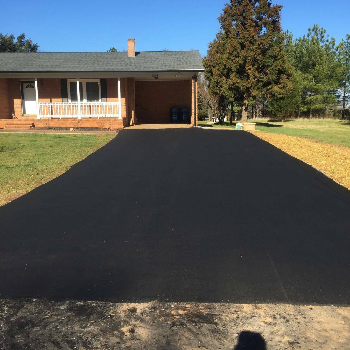 A newly paved asphalt driveway leading to a brick house with a porch, surrounded by grass and trees. Driveway Sealcoating Driveway Sealcoating