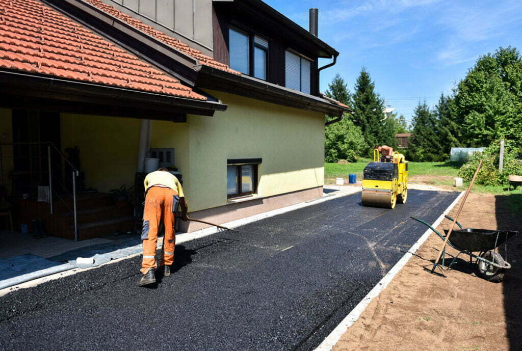 image-6-1-1024×689-1 Worker spreads asphalt on a driveway beside a yellow house; a steamroller is compacting the surface.
